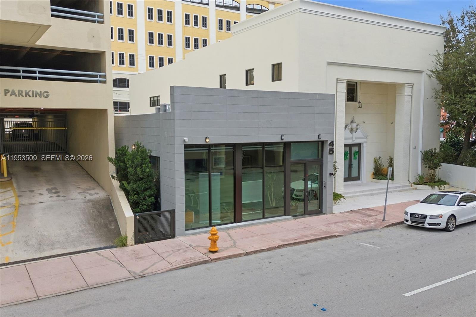 An exterior view of a gray, single-story commercial building situated between a parking garage and a white structure — 45 Valencia Ave, Coral Gables FL 33134, CORAL GABLES, FL