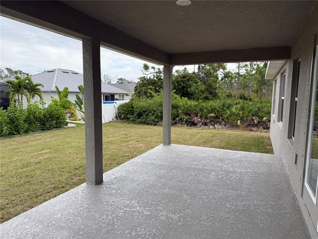 A covered concrete patio overlooks a grassy backyard with dense landscaping and neighboring houses — 14184 SALVATIERRA LANE, PORT CHARLOTTE, FL