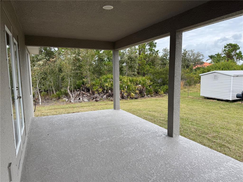 A covered concrete patio overlooks a grassy backyard with a shed and wooded area — 14184 SALVATIERRA LANE, PORT CHARLOTTE, FL