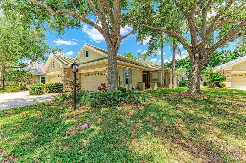 A tan single-story house with a two-car garage and large oak trees in the yard — 5047 LAKESCENE PLACE, SARASOTA, FL