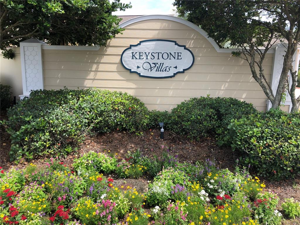 A beige sign for Keystone Villas is surrounded by green bushes and colorful flowers — 308 VIDALIA PLACE, THE VILLAGES, FL