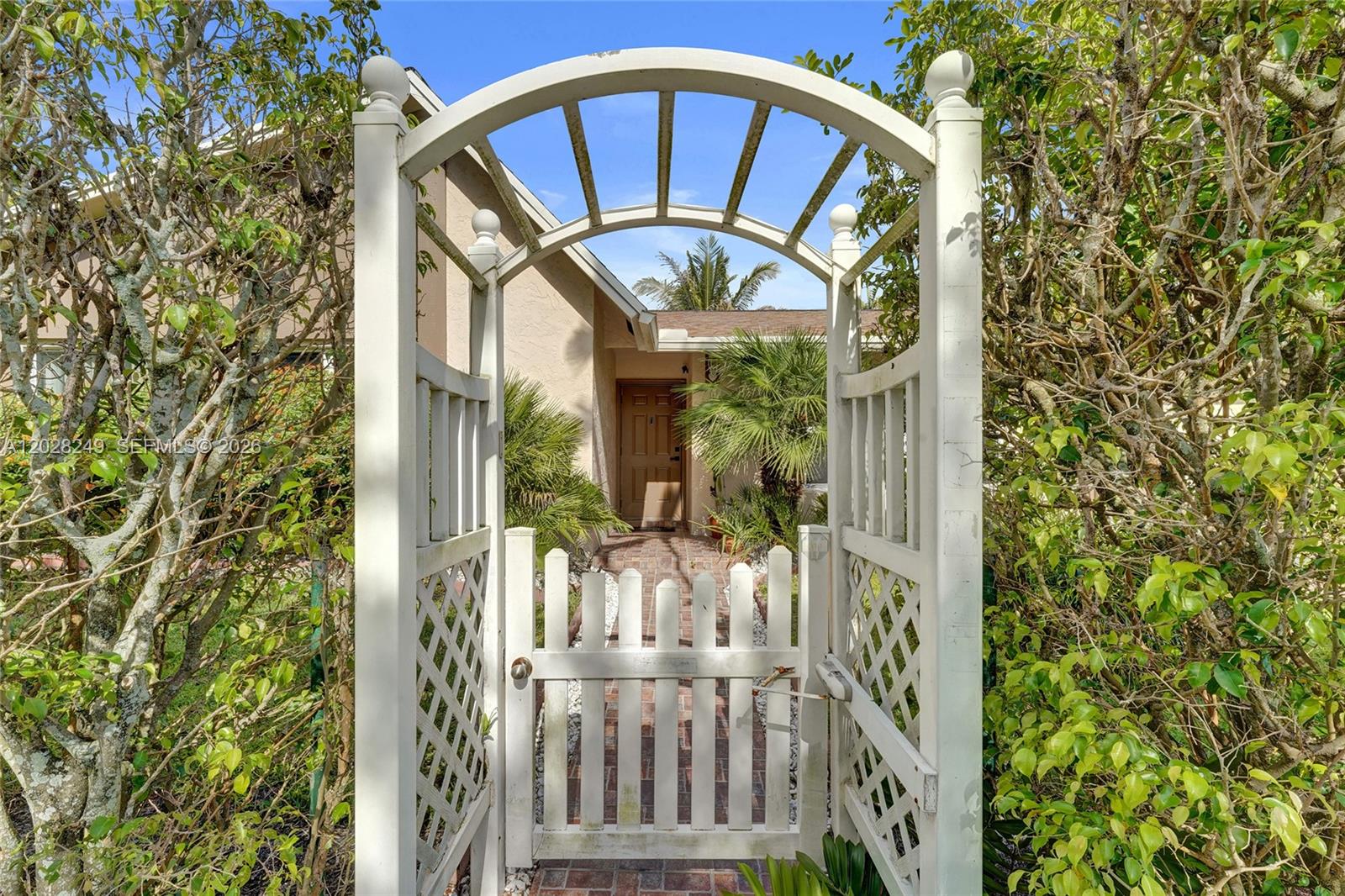 A white wooden arbor and gate lead to a brick walkway toward a house entrance — 417 SE 4th St, Dania Beach FL 33004, DANIA BEACH, FL