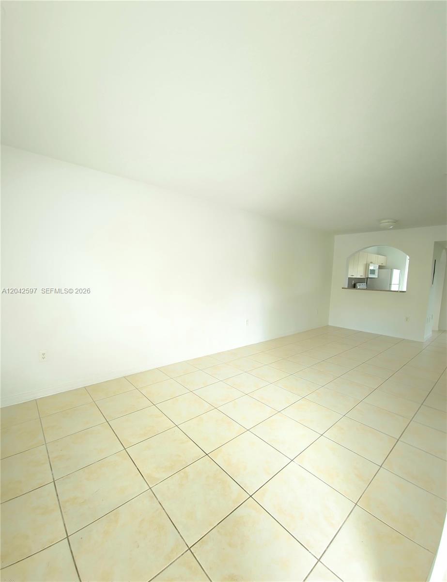 An empty living room features light-colored floor tiles and a view into the kitchen — 940 NE 33rd Ter # 102, Homestead FL 33033, HOMESTEAD, FL