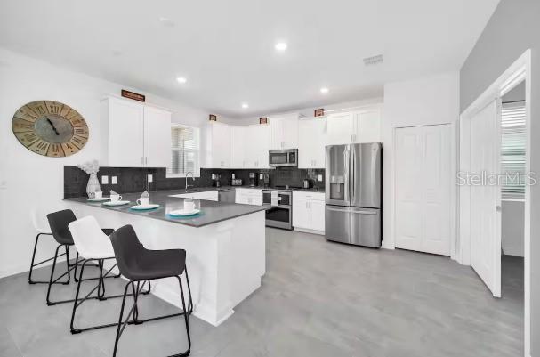 A modern kitchen features white cabinetry, stainless steel appliances, a breakfast bar, and gray flooring — 9092 SOMMERSET HILLS DRIVE, DAVENPORT, FL