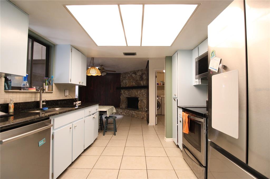 A galley-style kitchen features light blue cabinets, dark granite countertops, and a stone fireplace beyond — 1624 SW 77TH TERRACE, GAINESVILLE, FL