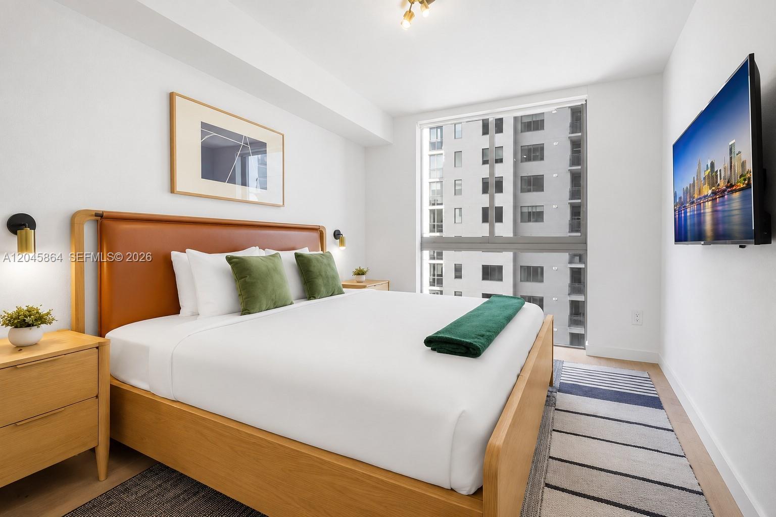 A modern bedroom features a wood-framed bed, green accents, and a large window overlooking buildings — 11 NE 6th St # 1712, Miami FL 33132, MIAMI, FL