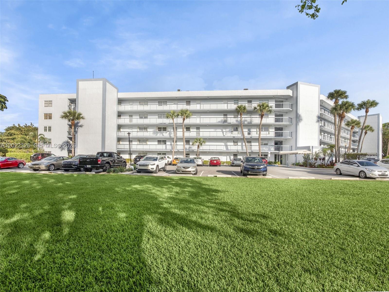A multi-story white apartment building stands behind a parking lot and a large grassy lawn — 6400 NW 2nd Ave # 117, Boca Raton FL 33487, BOCA RATON, FL