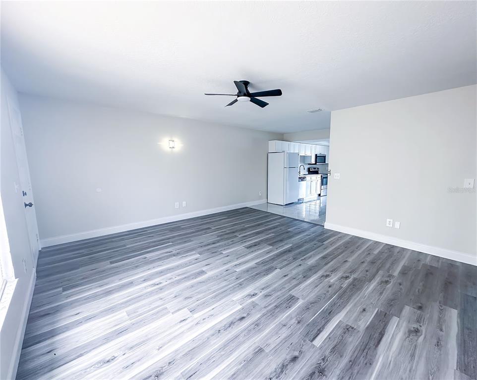 An empty living room with gray wood-look flooring and a ceiling fan leads to a kitchen — 15 W COUNTRY COVE, KISSIMMEE, FL