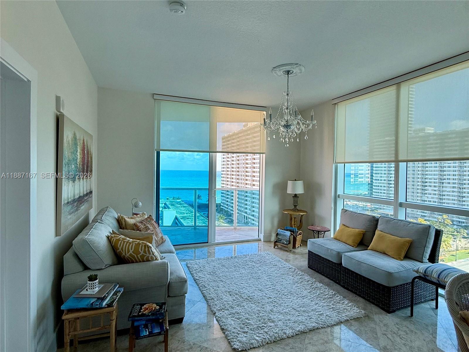 A living room features a chandelier, a white rug, and floor-to-ceiling windows overlooking the ocean — 1945 S Ocean Dr # 1201, Hallandale Beach FL 33009, HALLANDALE BEACH, FL