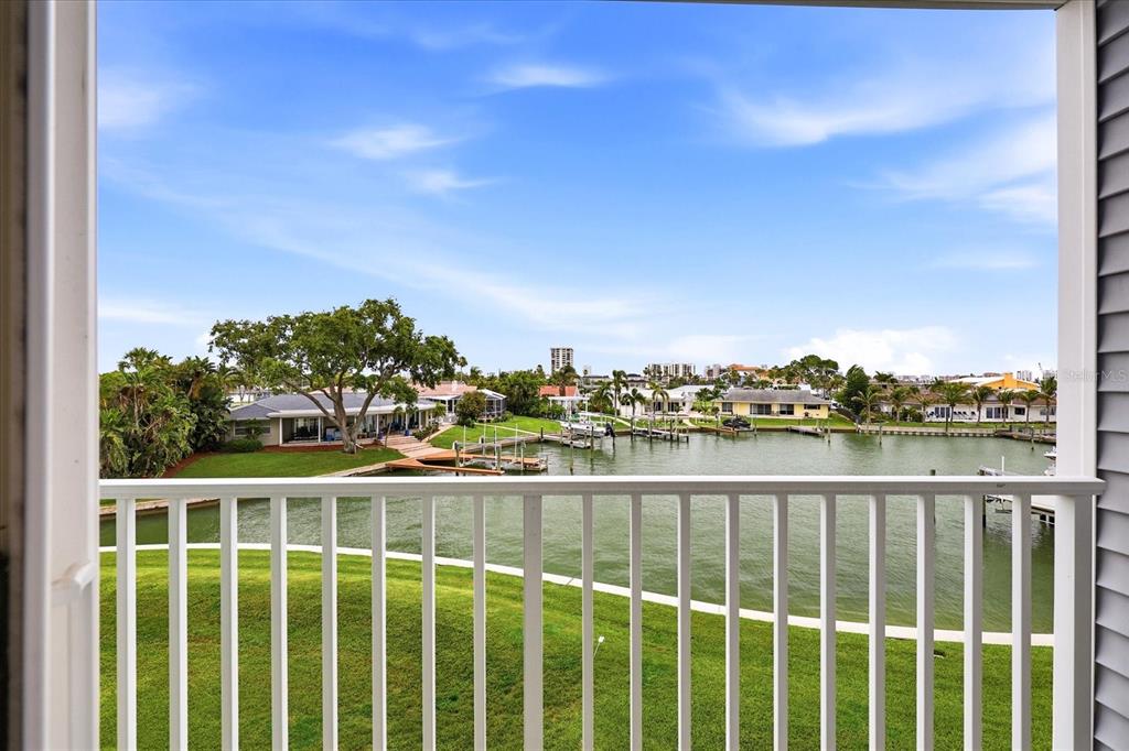 A balcony view overlooking a canal with boat docks, green lawns, and distant houses — 610 ISLAND WAY UNIT 201, CLEARWATER BEACH, FL