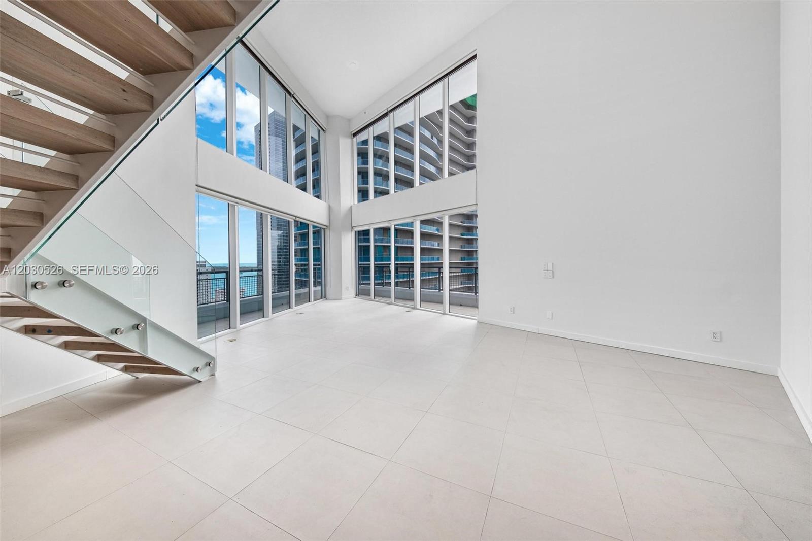 A modern, double-height living room features white tile floors, floor-to-ceiling windows, and a glass-railed staircase — 60 SW 13th St # 2600, Miami FL 33130, MIAMI, FL