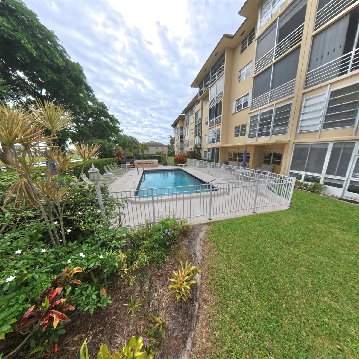 A rectangular swimming pool is enclosed by a white fence next to a multi-story building — 4100 Crystal Lake Drive # 209, Pompano Beach FL 33064, POMPANO BEACH, FL