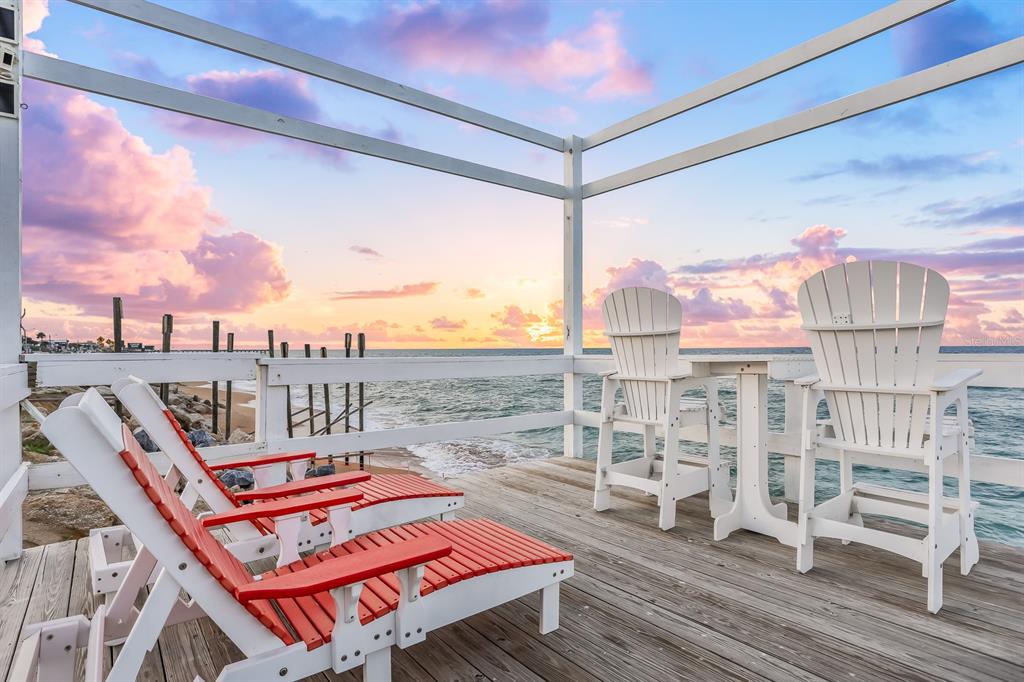 A wooden deck features red lounge chairs and white Adirondack chairs overlooking the ocean sunset — 1204 S OCEAN SHORE BOULEVARD, FLAGLER BEACH, FL