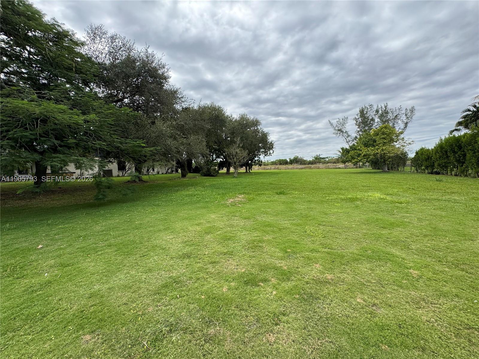 A large, grassy backyard features mature trees and a hedge under a cloudy sky — 18750 SW 207th Ave, Miami FL 33187, MIAMI, FL