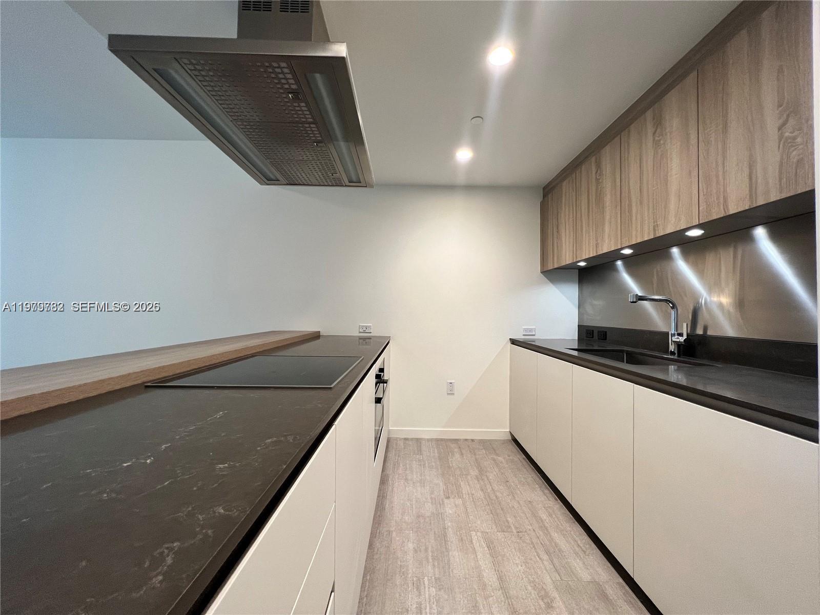 A modern kitchen features white cabinetry, dark countertops, a stainless steel range hood, and wood-toned upper cabinets — 1000 Brickell Plz # 3503, Miami FL 33131, MIAMI, FL