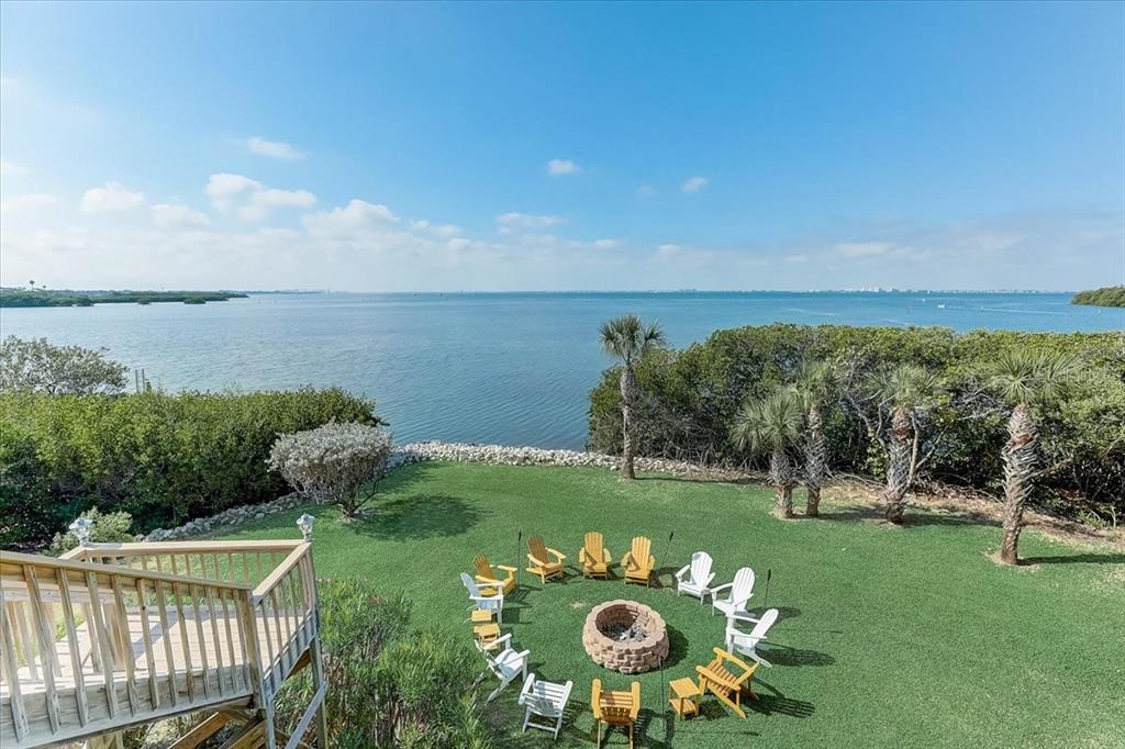 A grassy backyard features a circular fire pit surrounded by chairs overlooking a large bay — 2424 BAY DRIVE UNIT 2424/2426, BRADENTON, FL