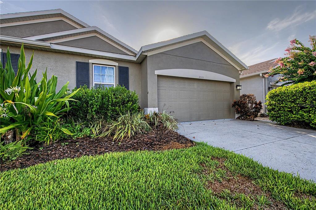 A single-story house exterior features a beige stucco facade, a two-car garage, and landscaped gardens — 10331 HERON HIDEAWAY LOOP, LAND O LAKES, FL