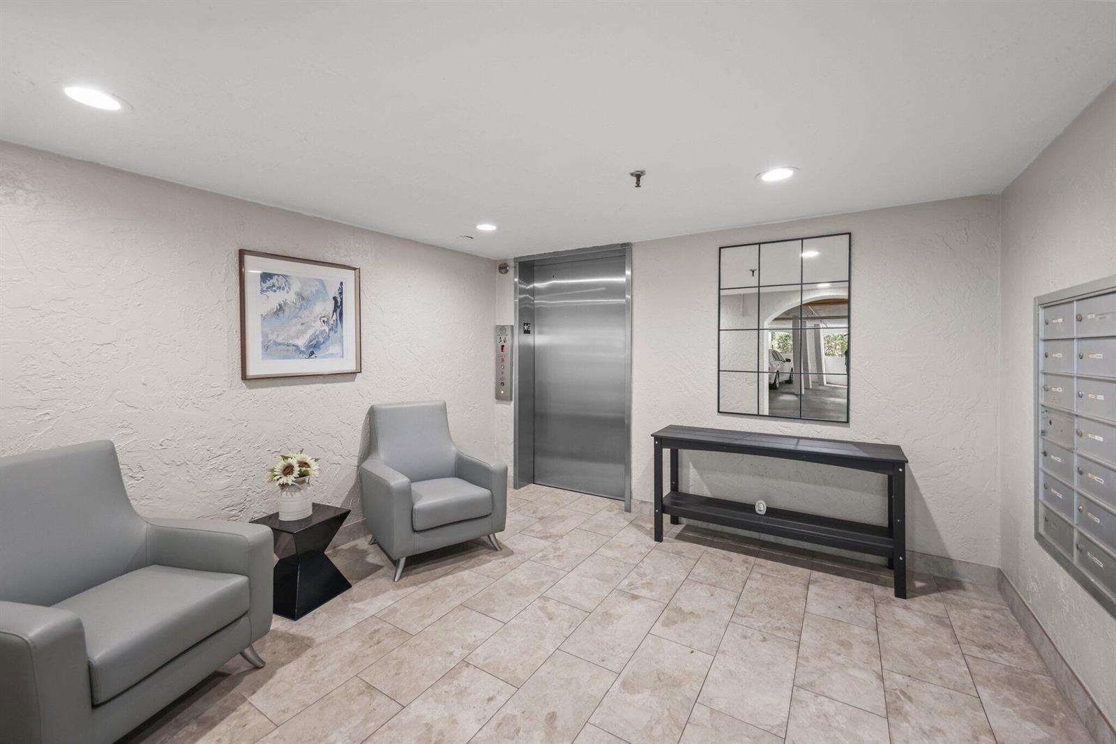 An apartment building lobby features two gray chairs, an elevator, a console table, and mailboxes — 7370 Orangewood Lane # 302, Boca Raton FL 33433, BOCA RATON, FL