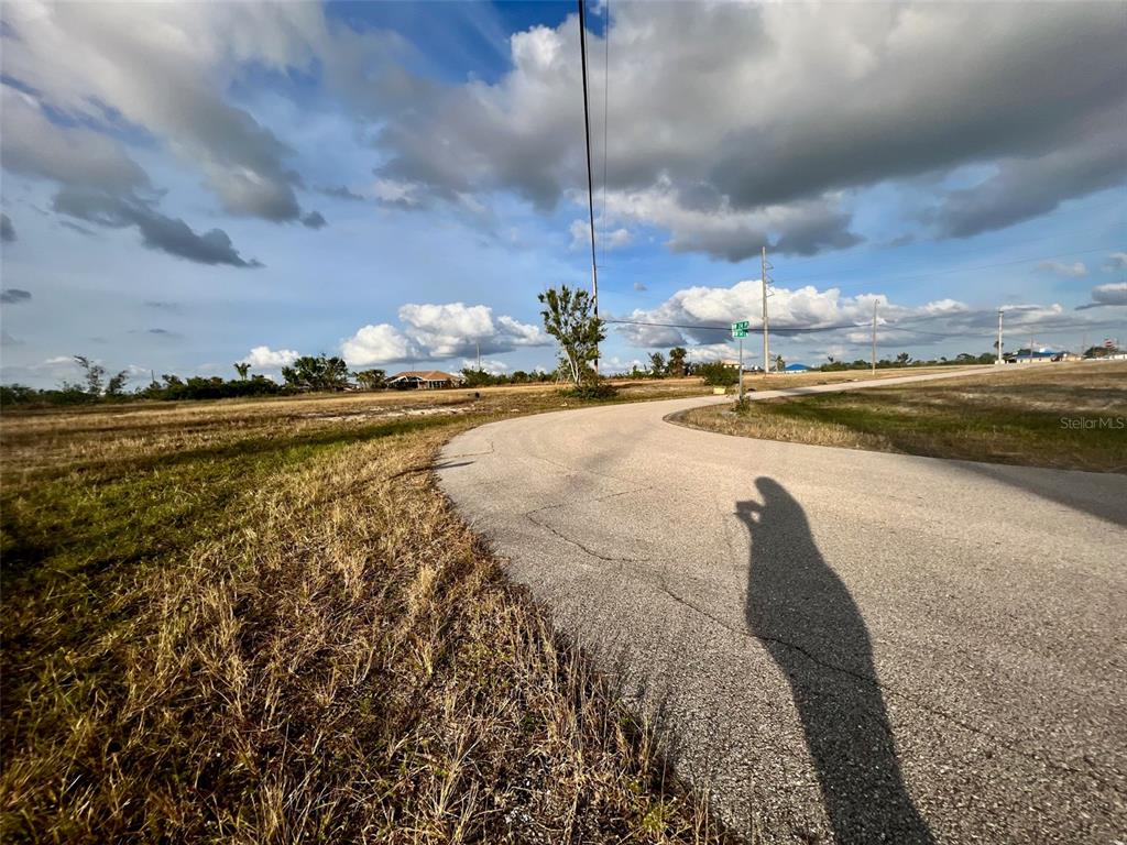 A paved road curves through a vacant, grassy lot under a cloudy blue sky — 1430 NW 24TH PLACE, CAPE CORAL, FL