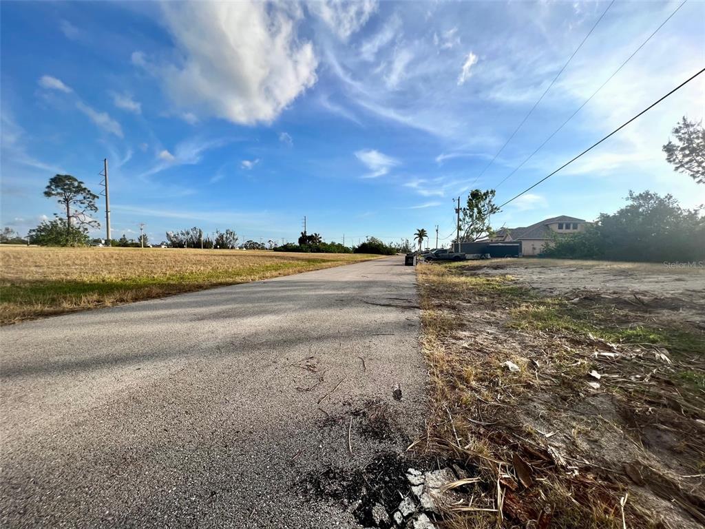 A paved road stretches through a grassy, undeveloped lot under a bright blue sky — 1430 NW 24TH PLACE, CAPE CORAL, FL