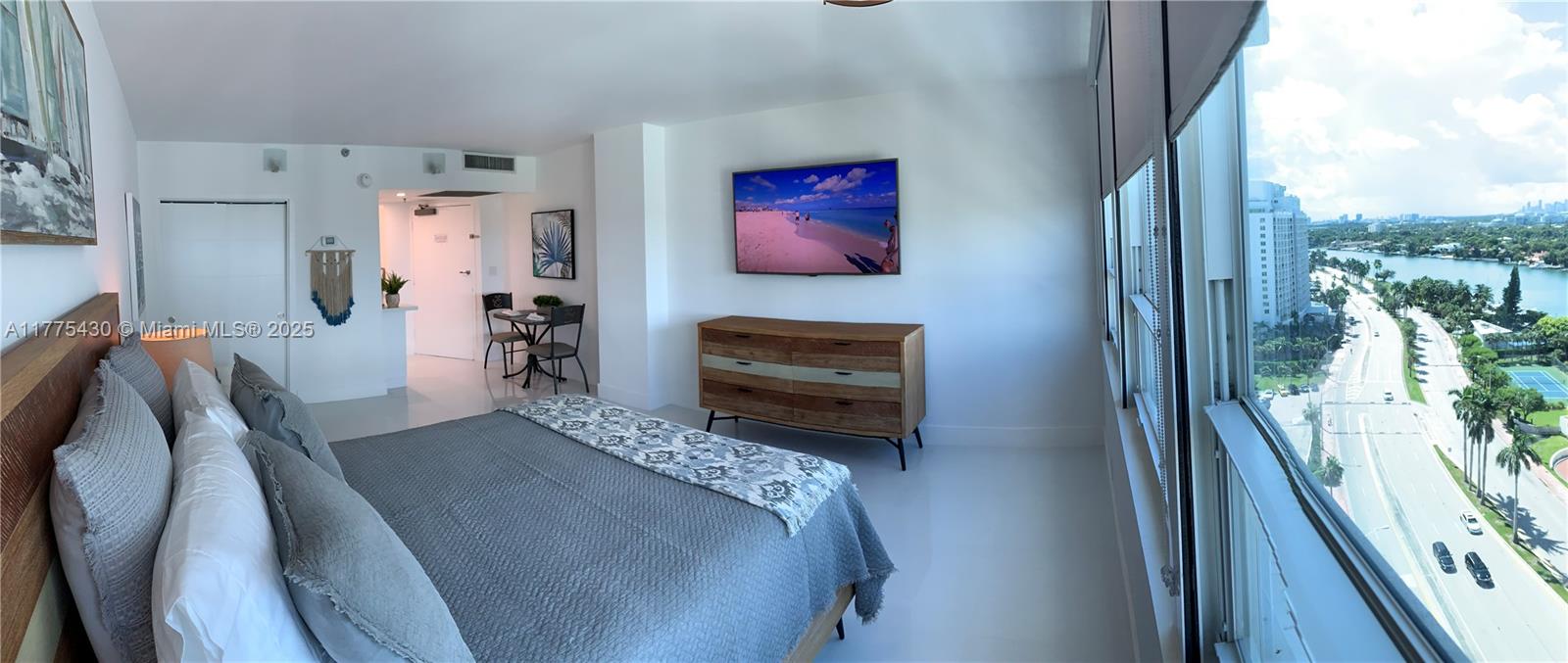 A bedroom features a bed, a wooden dresser, a wall-mounted television, and floor-to-ceiling windows — 5445 Collins Ave # 1520, Miami Beach FL 33140, MIAMI BEACH, FL