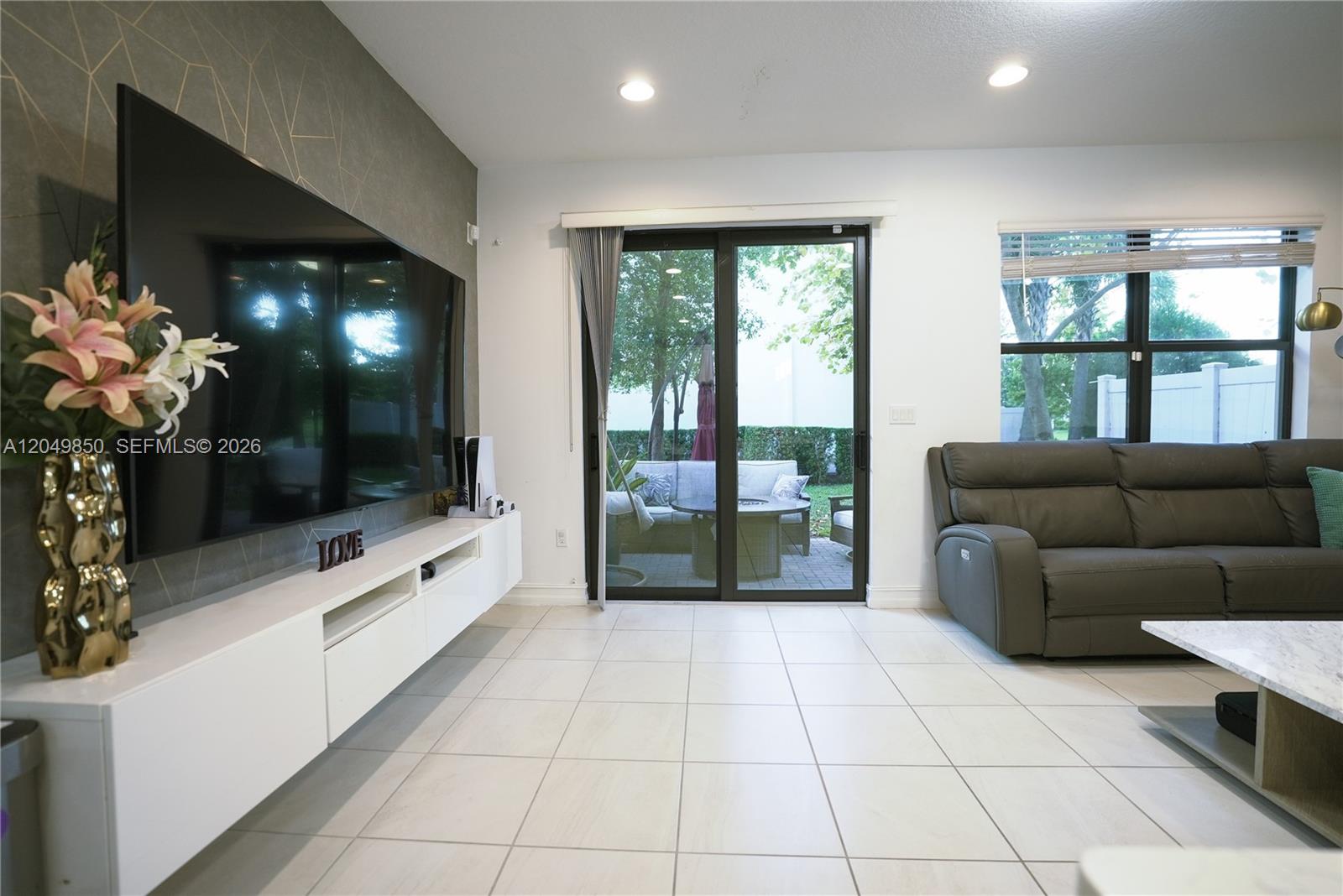 A living room features a wall-mounted television, white media console, tile flooring, and sliding glass doors — 10111 Akenside Dr, Boca Raton FL 33428, BOCA RATON, FL