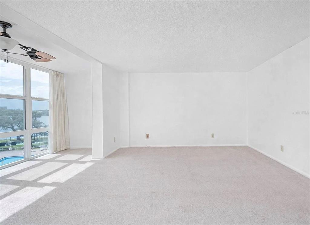 An empty living room features beige carpeting, white walls, and large windows overlooking a pool — 1224 S PENINSULA DRIVE UNIT 415, DAYTONA BEACH, FL