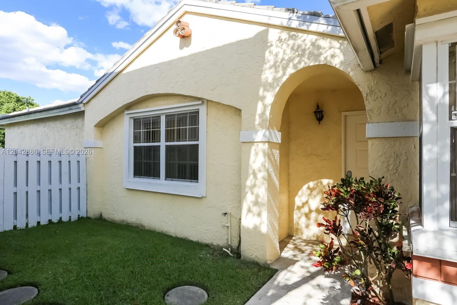 A yellow stucco house exterior features an arched entryway, a window, and a small lawn — PLANTATION, FL