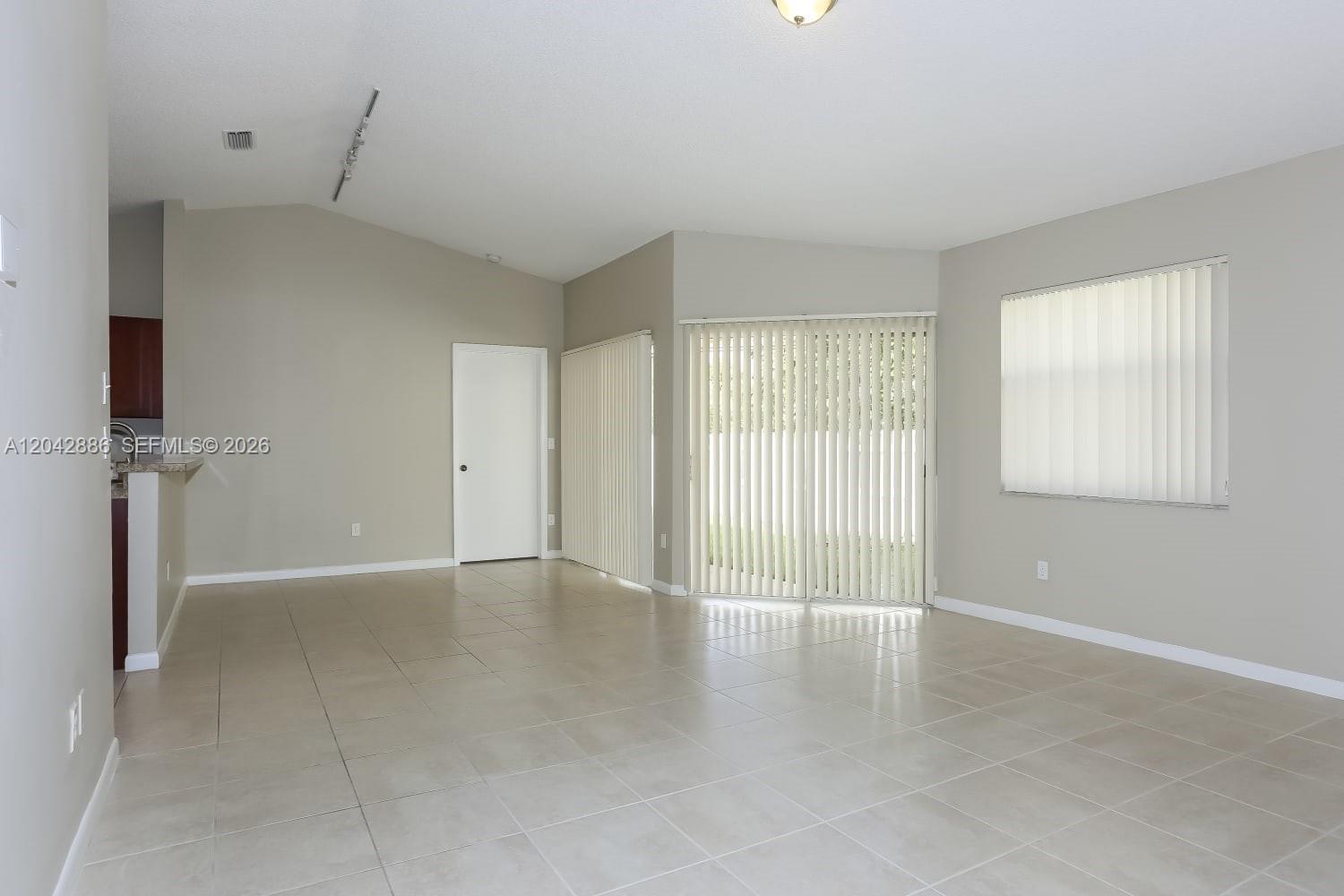 An empty living room features light-colored tile flooring, neutral walls, and vertical window blinds — PLANTATION, FL