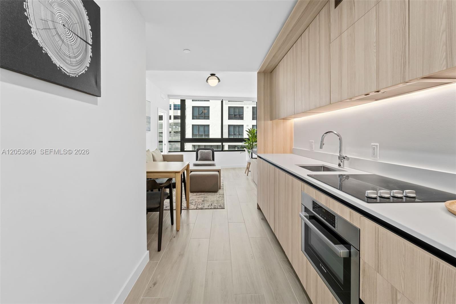 A modern kitchen with light wood cabinetry leads into a living area with large windows — 225 N Miami Ave # 912, Miami FL 33128, MIAMI, FL