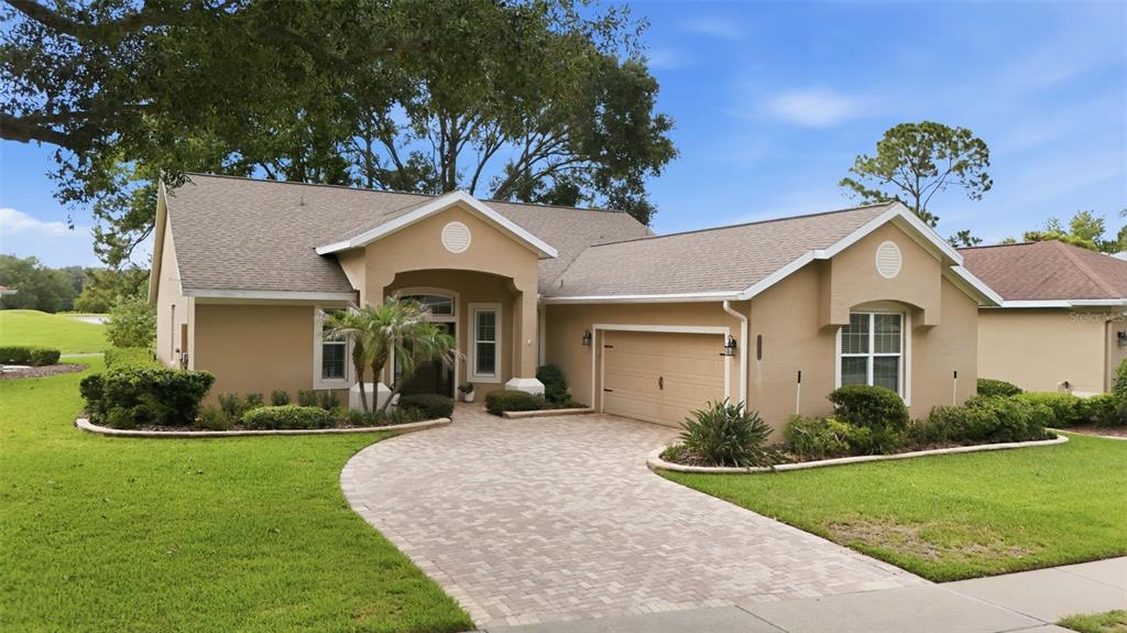 A tan, single-story house with a paver driveway, two-car garage, and a landscaped front yard — 7029 PINE HOLLOW DRIVE, MOUNT DORA, FL