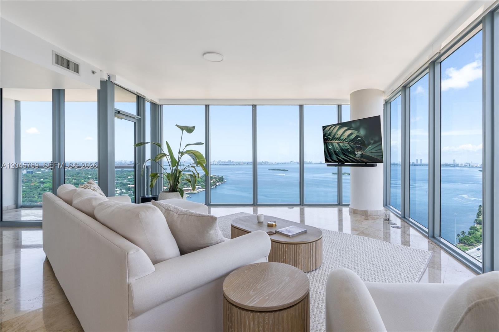 A modern living room features a white sofa, round coffee tables, and floor-to-ceiling windows overlooking water — 601 NE 36th St # 3112, Miami FL 33137, MIAMI, FL