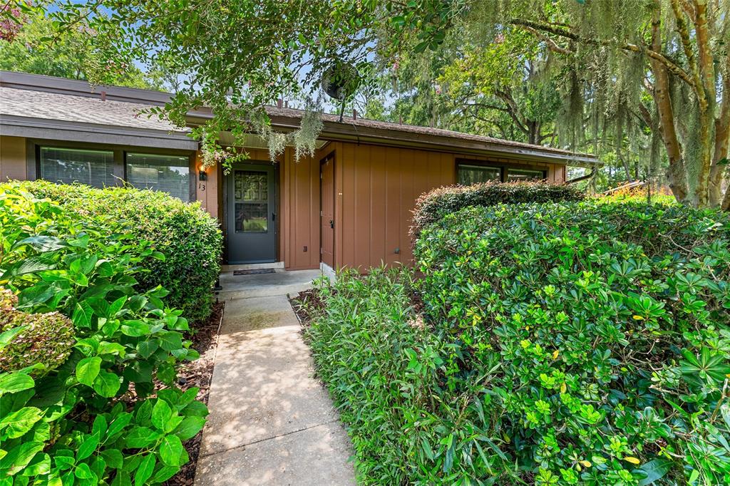 A concrete walkway leads to the front entrance of a brown house with dense landscaping — 19660 SW 83RD PLACE ROAD UNIT C13, DUNNELLON, FL