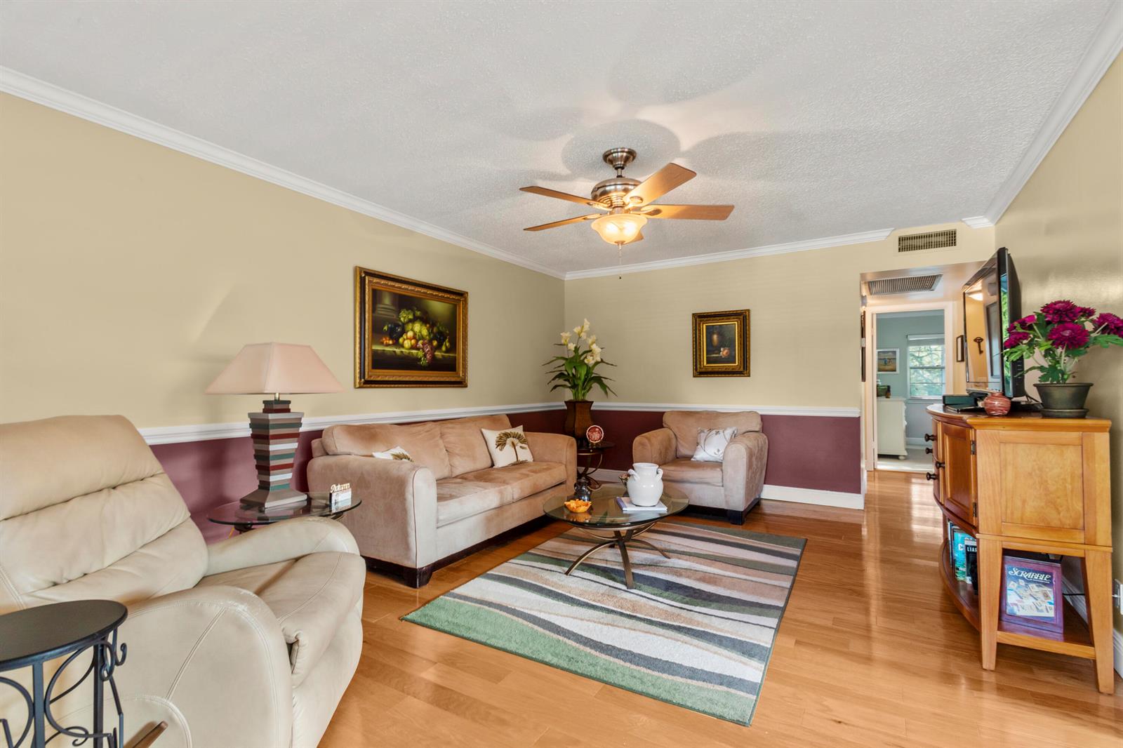 A living room features beige and maroon walls, hardwood floors, a ceiling fan, and furniture — 144 Tilford G Place # 144, Deerfield Beach FL 33442, DEERFIELD BEACH, FL