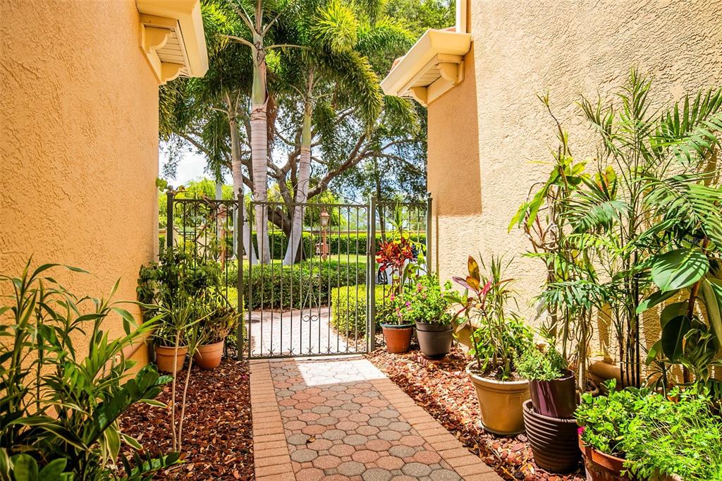 A paved walkway leads through a gated entrance between two stucco walls with potted plants — 3663 SQUARE WEST LANE UNIT 12, SARASOTA, FL