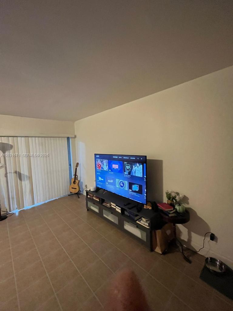 A living room features tile flooring, a television on a media console, and a guitar — 18111 NW 68th Ave # H102, Hialeah FL 33015, HIALEAH, FL