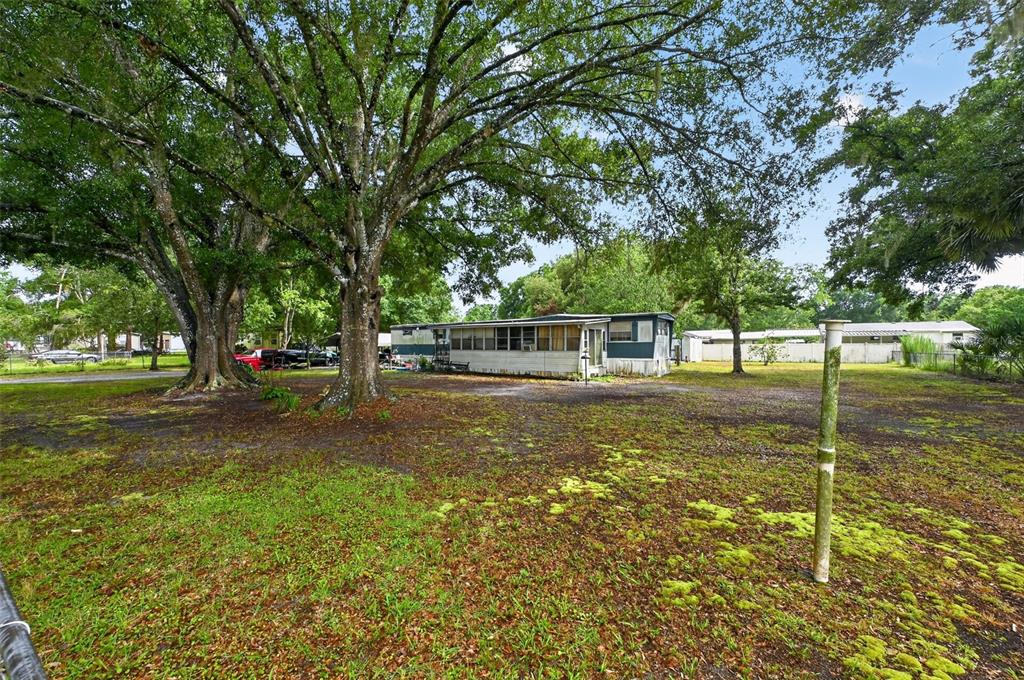 A mobile home sits on a grassy, tree-filled lot under a bright, cloudy sky — 26322 BRAHMA DRIVE, WESLEY CHAPEL, FL
