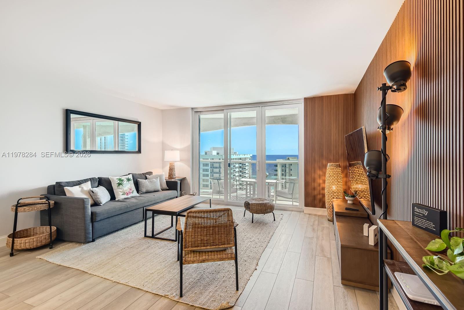 A living room features a gray sofa, wood-slat accent wall, and sliding doors overlooking the ocean — 3800 S Ocean Dr, Hollywood FL 33019, HOLLYWOOD, FL