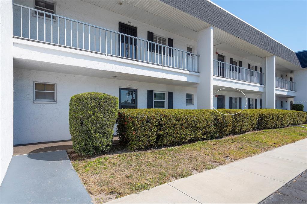 A two-story apartment building exterior features a ground-level walkway, white walls, and a hedge — 305 47TH AVENUE DRIVE W UNIT 146, BRADENTON, FL