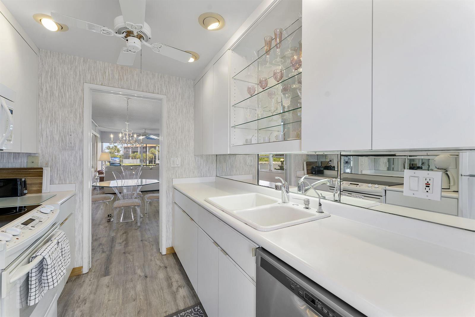 A white kitchen features a double sink, glass-front cabinets, and a mirrored backsplash — 101 NE 19th Avenue # 228, Deerfield Beach FL 33441, DEERFIELD BEACH, FL