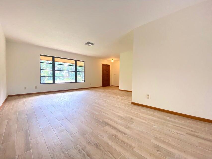 An empty living room features light wood-look tile flooring, white walls, and a large window — 5585 Pennock Point Road, Jupiter FL 33458, JUPITER, FL
