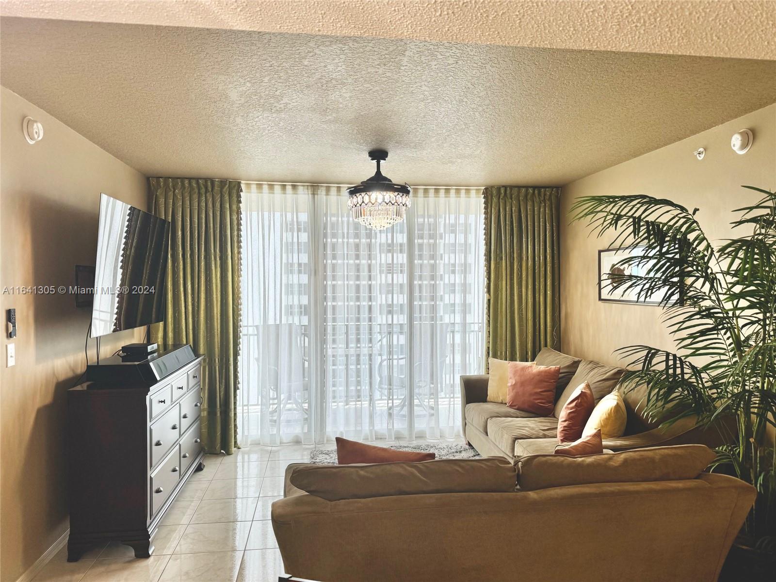 A living room features a sectional sofa, a television on a dresser, and a crystal chandelier — 2080 S Ocean Dr # 1607, Hallandale Beach FL 33009, HALLANDALE BEACH, FL