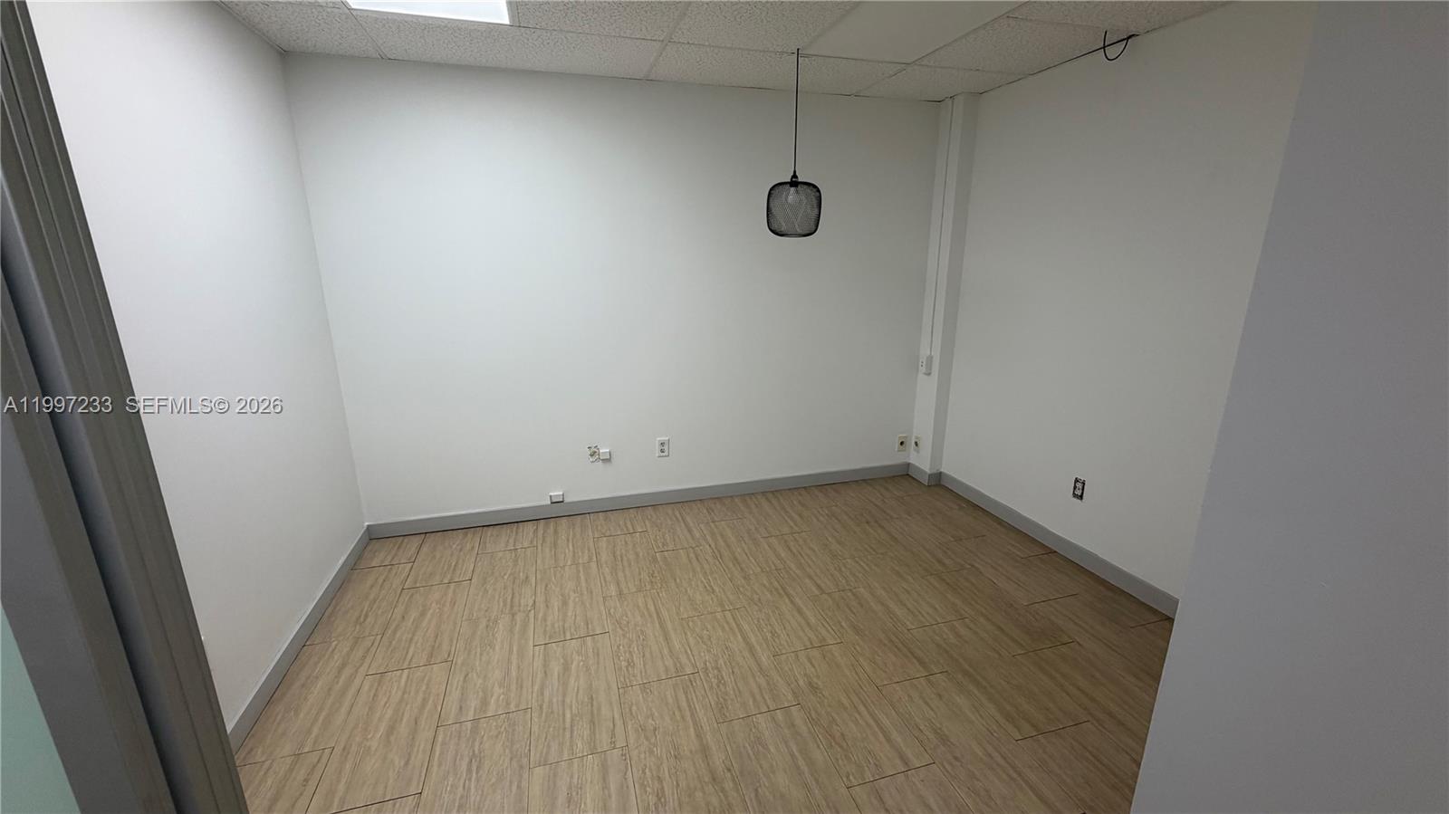 An empty office room features white walls, wood-look tile flooring, and a single hanging light — 10859 NW 29th St # 10859, Doral FL 33172, DORAL, FL