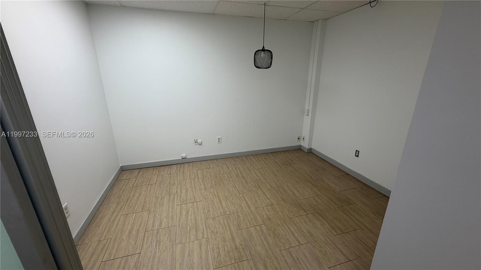 An empty office room features white walls, light wood-look tile flooring, and a hanging pendant light — 10859 NW 29th St # 10859, Doral FL 33172, DORAL, FL