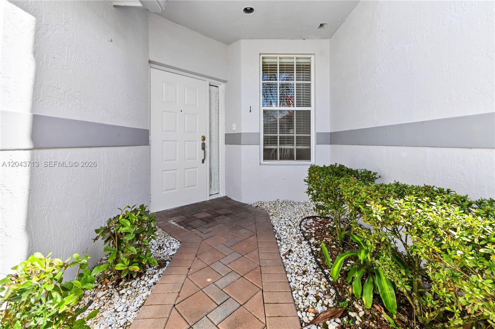 A white front door with a side window leads to a brick walkway and landscaping — 7874 Laina Ln # 1, Boynton Beach FL 33437, BOYNTON BEACH, FL