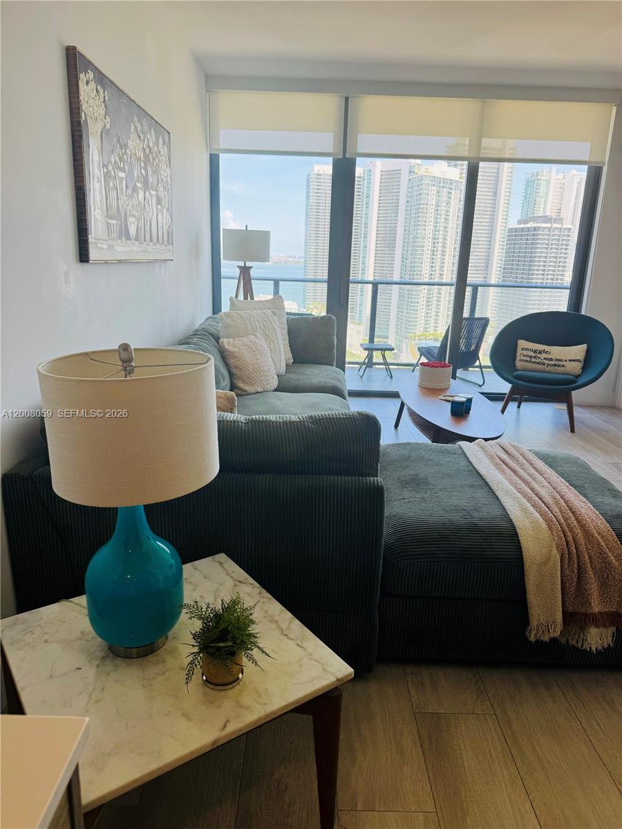 A living room features a green sectional sofa, a teal lamp, and floor-to-ceiling windows overlooking skyscrapers — 121 NE 34th St # 2705, Miami FL 33137, MIAMI, FL