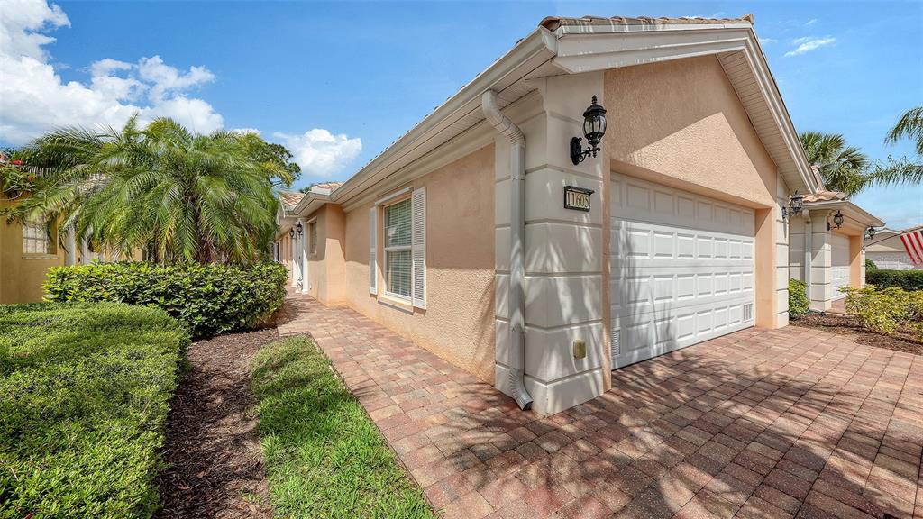 A peach-colored house features a white garage door, a brick driveway, and lush landscaping — 11605 GARESSIO LANE, SARASOTA, FL