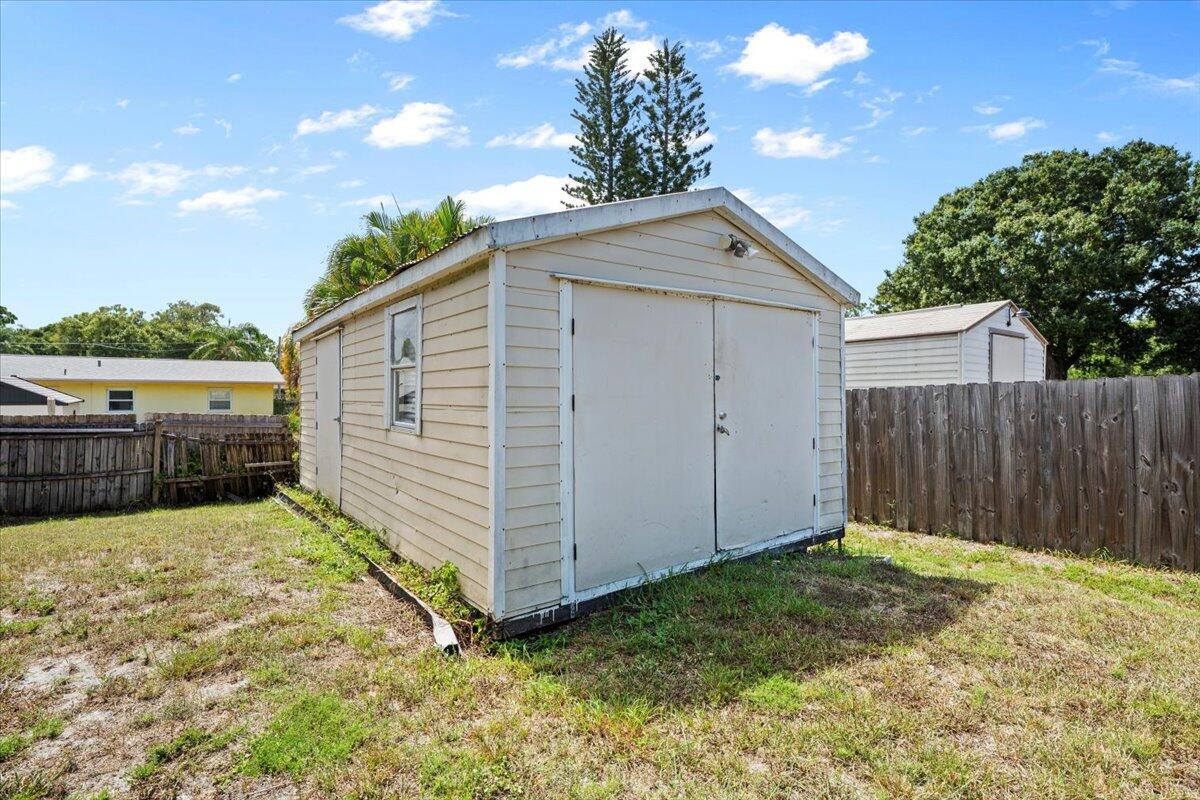 A beige storage shed with double doors stands in a grassy backyard near a wooden fence — 6015 Raintree Trl, Fort Pierce FL 34982, FORT PIERCE, FL