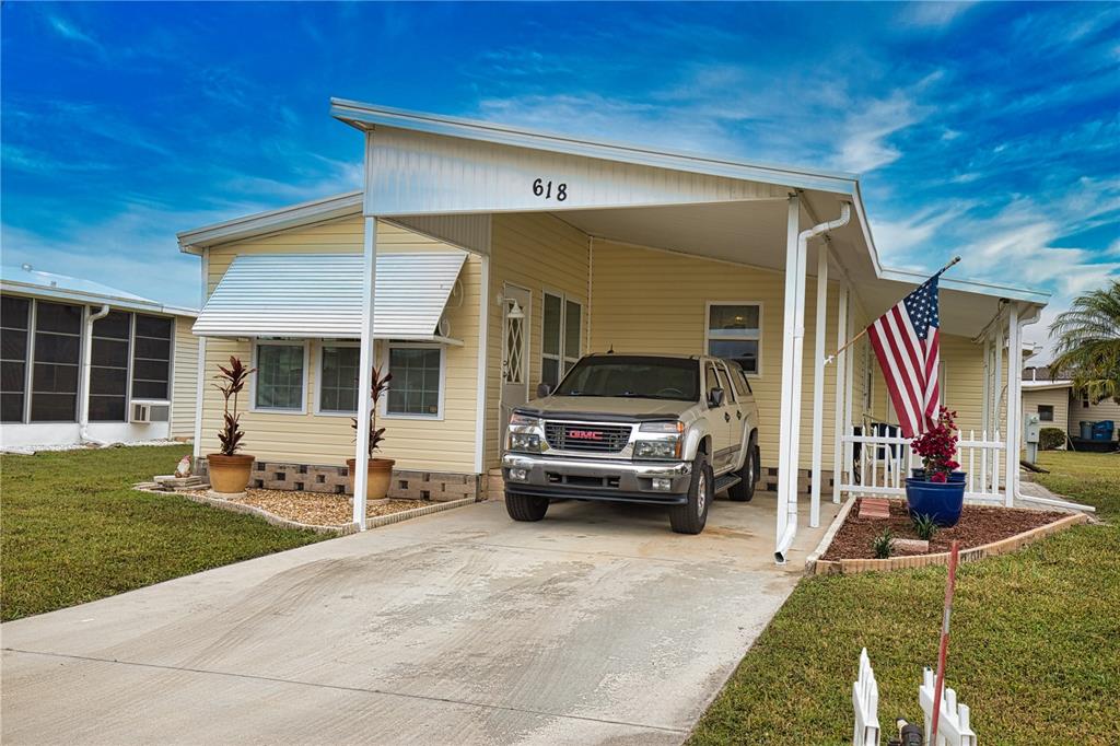 A light yellow manufactured home with a carport and an American flag on the porch — 618 IGLESIA DRIVE, NORTH PORT, FL