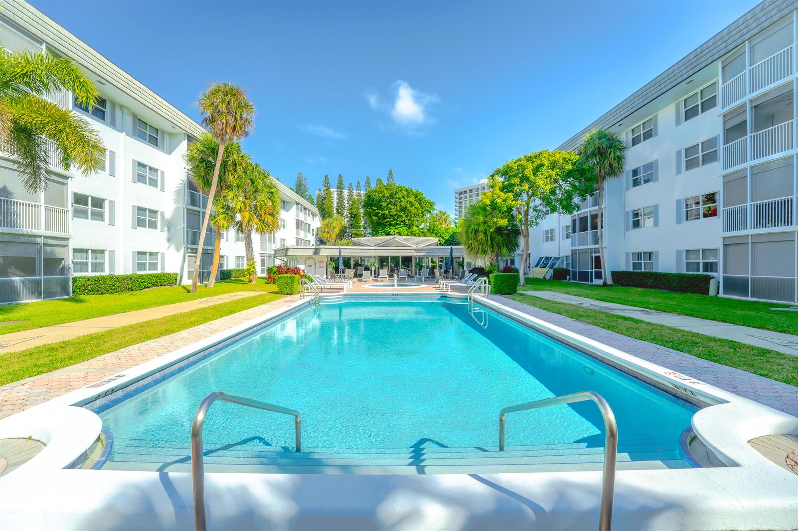 A rectangular swimming pool sits between two white multi-story apartment buildings with palm trees — 800 E Camino Real # 309, Boca Raton FL 33432, BOCA RATON, FL